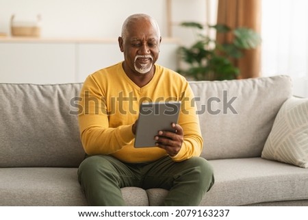 Senior Black Man Using Digital Tablet Browsing Internet Sitting On Couch At Home. Mature Male Watching Film Online On Computer Relaxing In Living Room On Weekend. Older People And Gadgets Stock foto © 