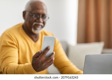 Senior Black Man Using Phone Wearing Eyeglasses Sitting At Laptop On Sofa Indoors. Selective Focus On Smartphone While Male Using Mobile Application. People And Gadgets, Eyesight Concept