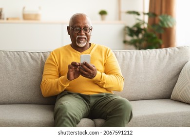 Senior Black Man mit Smartphone Texting Networking Online und Scrollen News in Social Media-Anwendungen auf Couch zu Hause sitzend. Reife männliche Lesemann Nachricht auf dem Telefon. Neues Konzept für mobile App
