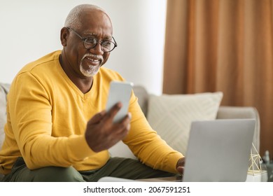Senior Black Man Using Smartphone And Laptop Computer Working Online Sitting On Sofa Indoors. Senior Male Professional Texting Via Cellphone And Browsing Internet In Modern Office. Selective Focus