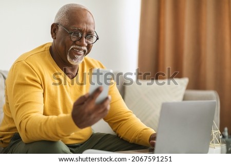Senior Black Man Using Smartphone And Laptop Computer Working Online Sitting On Sofa Indoors. Senior Male Professional Texting Via Cellphone And Browsing Internet In Modern Office. Selective Focus Stock foto © 