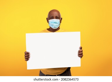 Senior Black Man Wearing Anti Covid Face Mask, Showing Empty Advertisement Board Over Orange Studio Background, Mockup. Elderly African American Male With Blank Poster Promoting Your Service