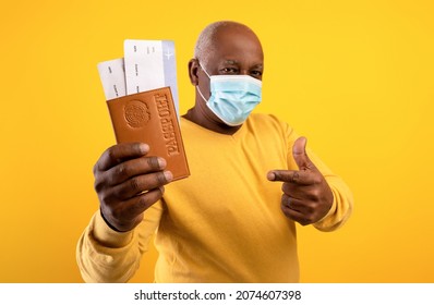 Senior Black Man Wearing Medical Mask, Pointing At Plane Tickets And Passport On Orange Studio Background. Elderly African American Male Traveling Abroad During Coronavirus Pandemic