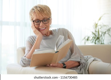 Senior blond woman in glasses resting on sofa and reading a book