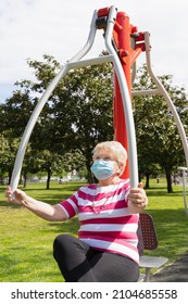 Senior Blonde Lady Doing Physical Activity At Park Exercise Machine With Face Mask And Eyeglasses. Elder Woman Training Upper Body To Keep Fit And Healthy On Sunny Day Outdoors