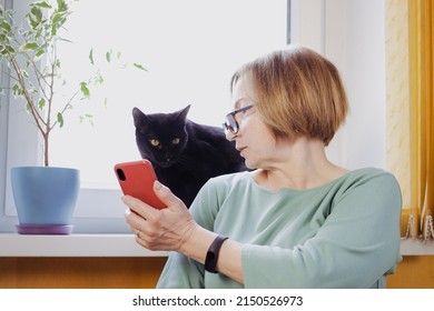 Senior Blonde Woman Using Mobile Phone For Video Call With Friend Or Children, Showing Black Pet Cat, Sitting Near Window At Home. Online Consultation With The Veterinarian Using Wireless 5g Internet