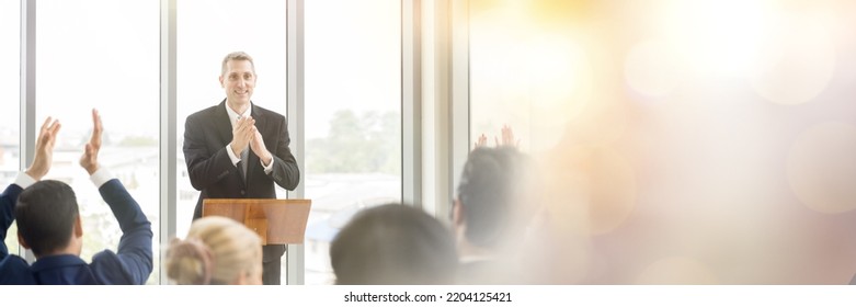 Senior Business Man Talking And Clapping Hands At Podium Speaker. Back View Of Business People Clapping Hands To Speaker At Business Conference Room. Banner, Bokeh And Light Concept