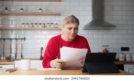 Senior business woman sit in kitchen using tablet device, check information on document, taking notes, analyze data, looking through papers busy in paperwork - Powered by Shutterstock