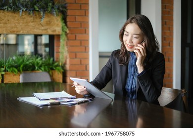 A Senior Business Woman Sitting At Desk And Holding And Looking At Tablet, While Calling With Someone With Smile At Co Work Space.