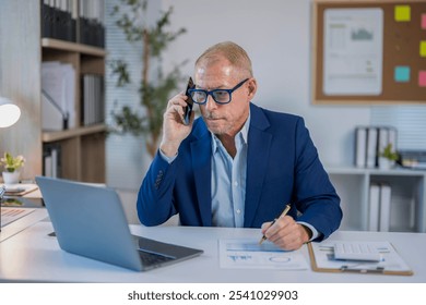 Senior businessman in blue suit working in modern office, talking on mobile phone and reviewing financial report on laptop - Powered by Shutterstock