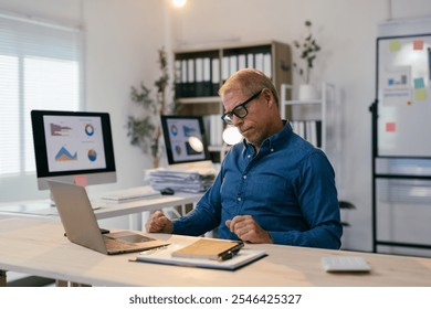 Senior businessman clenching his fists while reading upsetting news on his laptop in a modern office, surrounded by computers, documents, and charts, expressing frustration and stress - Powered by Shutterstock