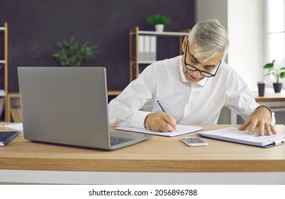 Senior Businessman Doing Paperwork And Taking Notes In His Business Planner Or Notebook. Busy Older Adult Man Sitting At Office Desk With Modern Laptop And Writing Something Down On Sheet Of Paper