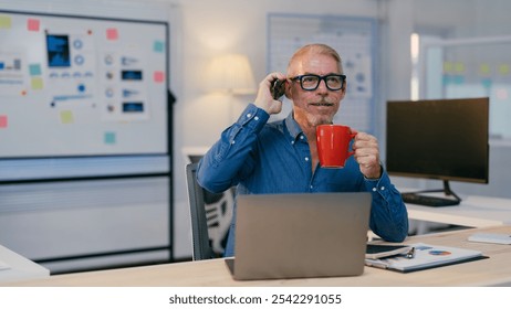 Senior businessman drinking from a red mug and talking on his mobile phone while working on a laptop in a modern office with charts and graphs on a whiteboard - Powered by Shutterstock