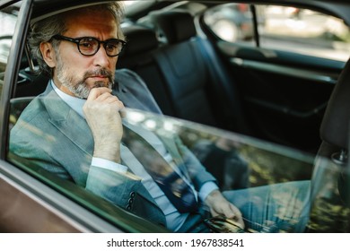 Senior Businessman Driving On Car Back Seat, Looking Away While Holding A Smart Phone.