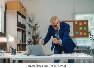 Senior businessman expressing excitement and joy while working on a laptop, celebrating a successful outcome in his modern office - Powered by Shutterstock