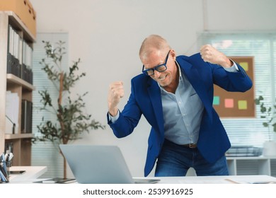 Senior businessman expressing excitement and joy while looking at his laptop in the office, celebrating a successful outcome - Powered by Shutterstock