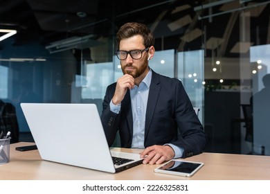 Senior businessman in glasses and beard is thinking about a decision, man is working inside the office and using laptop at work, mature investor in a business suit is sitting at the table - Powered by Shutterstock