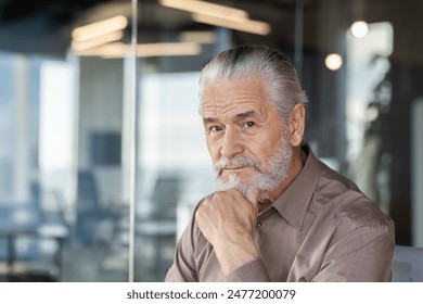 Senior businessman with grey hair and beard in a modern office setting, contemplating while looking at camera. Professional and thoughtful expression. - Powered by Shutterstock