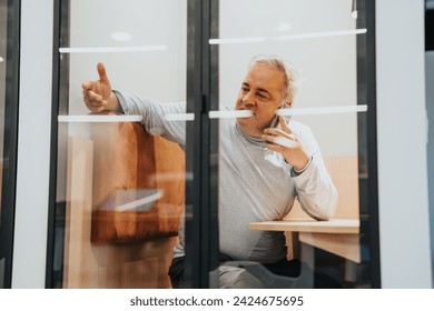 Senior businessman having phone call while sitting at soundproof phone booth.
