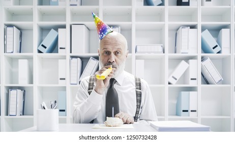 Senior Businessman Having A Sad Lonely Birthday In The Office, He Is Wearing A Party Hat And Having A Small Cake Alone