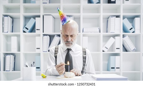 Senior Businessman Having A Sad Lonely Birthday In The Office, He Is Wearing A Party Hat And Having A Small Cake Alone