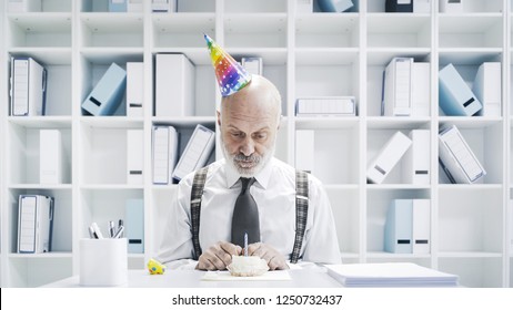 Senior Businessman Having A Sad Lonely Birthday In The Office, He Is Wearing A Party Hat And Having A Small Cake Alone