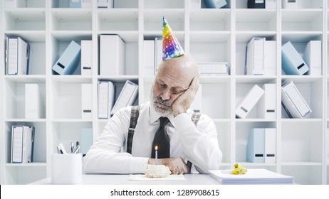 Senior Businessman Having A Sad Lonely Birthday In The Office, He Is Wearing A Party Hat And Having A Small Cake Alone