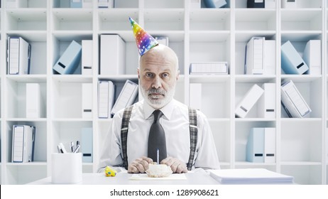 Senior Businessman Having A Sad Lonely Birthday In The Office, He Is Wearing A Party Hat And Having A Small Cake Alone