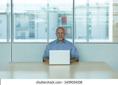 Senior Businessman With Laptop Sat At Desk In Office, Window In Background.