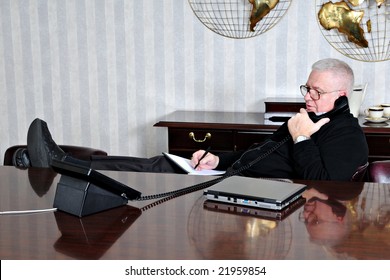 A senior businessman relaxed with his feet up while taking notes from a business phone conversation in the conference room.
