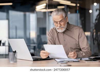 Senior businessman reviewing documents while sitting at a desk with a laptop in a modern office setting. - Powered by Shutterstock