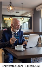 Senior Businessman Sitting At A Restaurant Table, Purchasing Over The Internet And Paying With A Credit Card