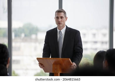 Senior Businessman Standing At Podium Giving A Speech Addressing A Conference In Seminar. Old Executive Man Presentation In Meeting At Office On Window With Colleagues	