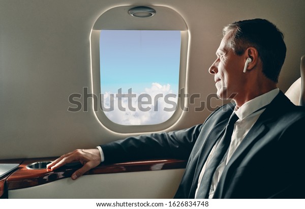 Senior businessman in suit flying\
in private jet and listening to music via wireless\
earphones.