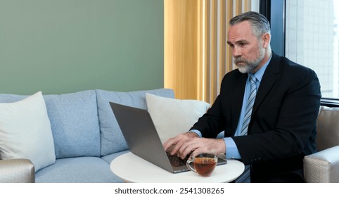 Senior businessman in suit and necktie typing on laptop computer while sitting in cafeteria during coffee break. A cup of hot tea are on the table. - Powered by Shutterstock