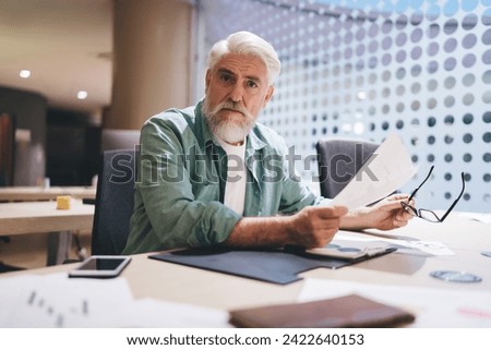 Senior businessman in teal shirt, holding glasses, scrutinizes a document at his office desk. Concern and attention to detail are evident in his expression.