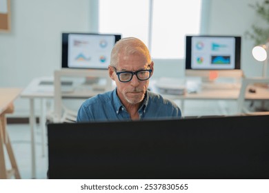 Senior businessman wearing glasses focusing on computer screen with charts displayed on other monitors in bright modern office, indicating dedicated work and data analysis - Powered by Shutterstock