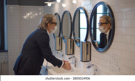 Senior businessman wearing safety mask washing hands in public restroom. Elegant confident entrepreneur in protective mask cleaning hands in sink after visiting office toilet