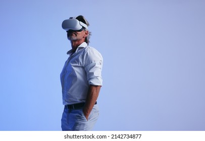 Senior businessman wearing a virtual reality headset in a studio. Mature businessman looking away while standing with his hands in his pockets. Businessman attending a virtual meeting in the metaverse - Shutterstock ID 2142734877