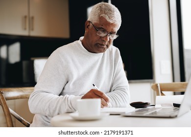 Senior businessman writing in a notebook while sitting at a desk in his home. Mature businessman making plans in his journal while doing remote freelance work. - Powered by Shutterstock