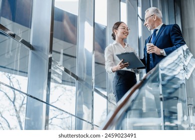 Senior businessman and young asian female working with documents together in office lobby. Smiling busy middle aged business man and woman standing and discussing digital strategy - Powered by Shutterstock