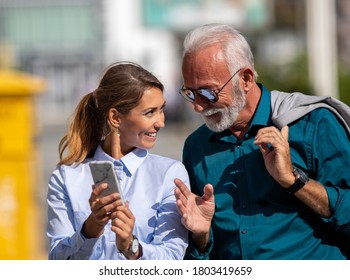 Senior businessman and young woman walking on street, smiling and looking at mobile phone