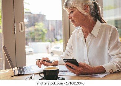 Senior businesswoman holding a smart phone sitting at coffee shop and working on laptop. Mature female entrepreneur at cafe using laptop computer. - Powered by Shutterstock