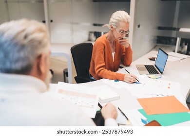 Senior businesswoman in orange cardigan focusing on data analysis, reviewing charts and graphs on paper. Open laptop and scattered documents on desk suggest a collaborative office environment - Powered by Shutterstock