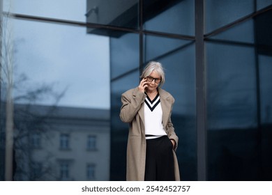 A senior businesswoman in professional attire talking on her phone outside a modern office building during daytime - Powered by Shutterstock