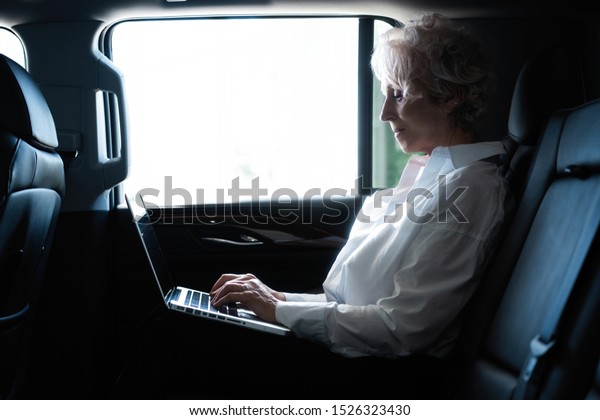 Senior businesswoman travelling to\
office in a car sitting on backseat with laptop. Businesswoman with\
laptop receiving a mail on the backseat of a\
car.