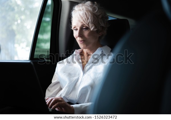 Senior businesswoman travelling to\
office in a car sitting on backseat with laptop. Businesswoman with\
laptop receiving a mail on the backseat of a\
car.