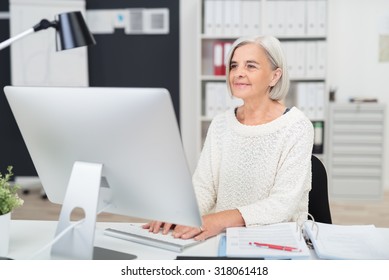 Senior Businesswoman At Work In The Office Seated At Her Desk Typing In Information On The Desktop Computer