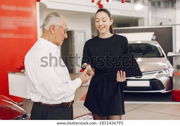 Senior in a car salon. Old man buying the car.\
Elegant woman helps a man buy a\
car.