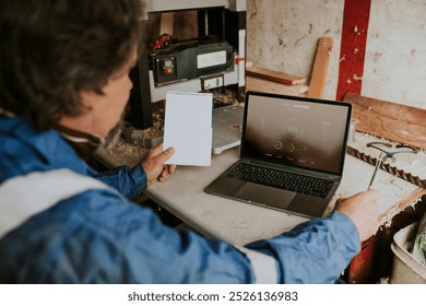 Senior carpenter man holding a white notebook while looking at his laptop on the desk, small business concept. Carpenter workshop. Middle age carpenter man working in his wood studio - Powered by Shutterstock